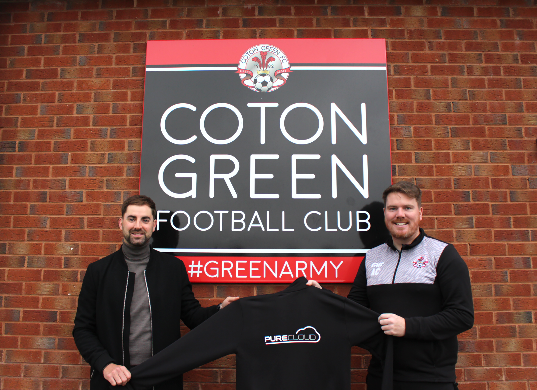 Two men holding a black sweatshirt with the PureCloud logo in front of the Coton Green Football Club sign.