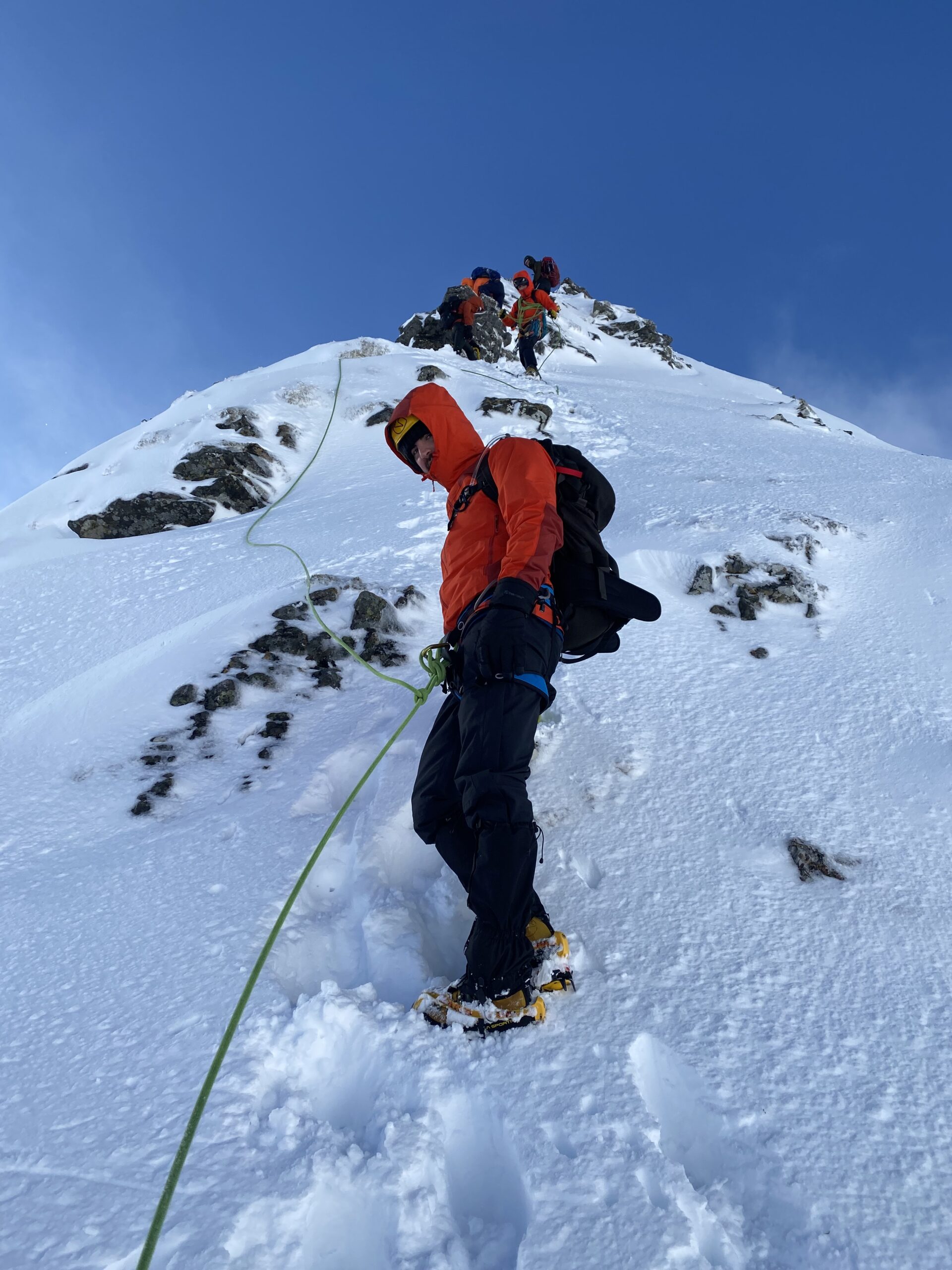 Climber in an orange jacket ascending a snowy mountain, secured by a rope, with other climbers visible in the background.