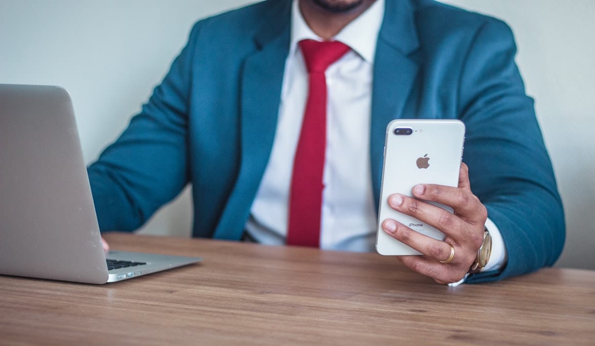 Man in a suit holding an iPhone while looking at his laptop.